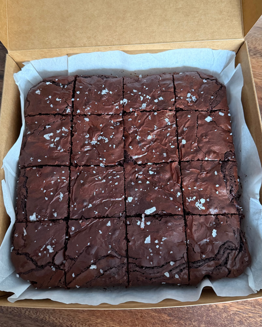 A box of chocolate brownies cut into a grid, topped with coarse sea salt flakes, presented in a cardboard box lined with parchment paper.